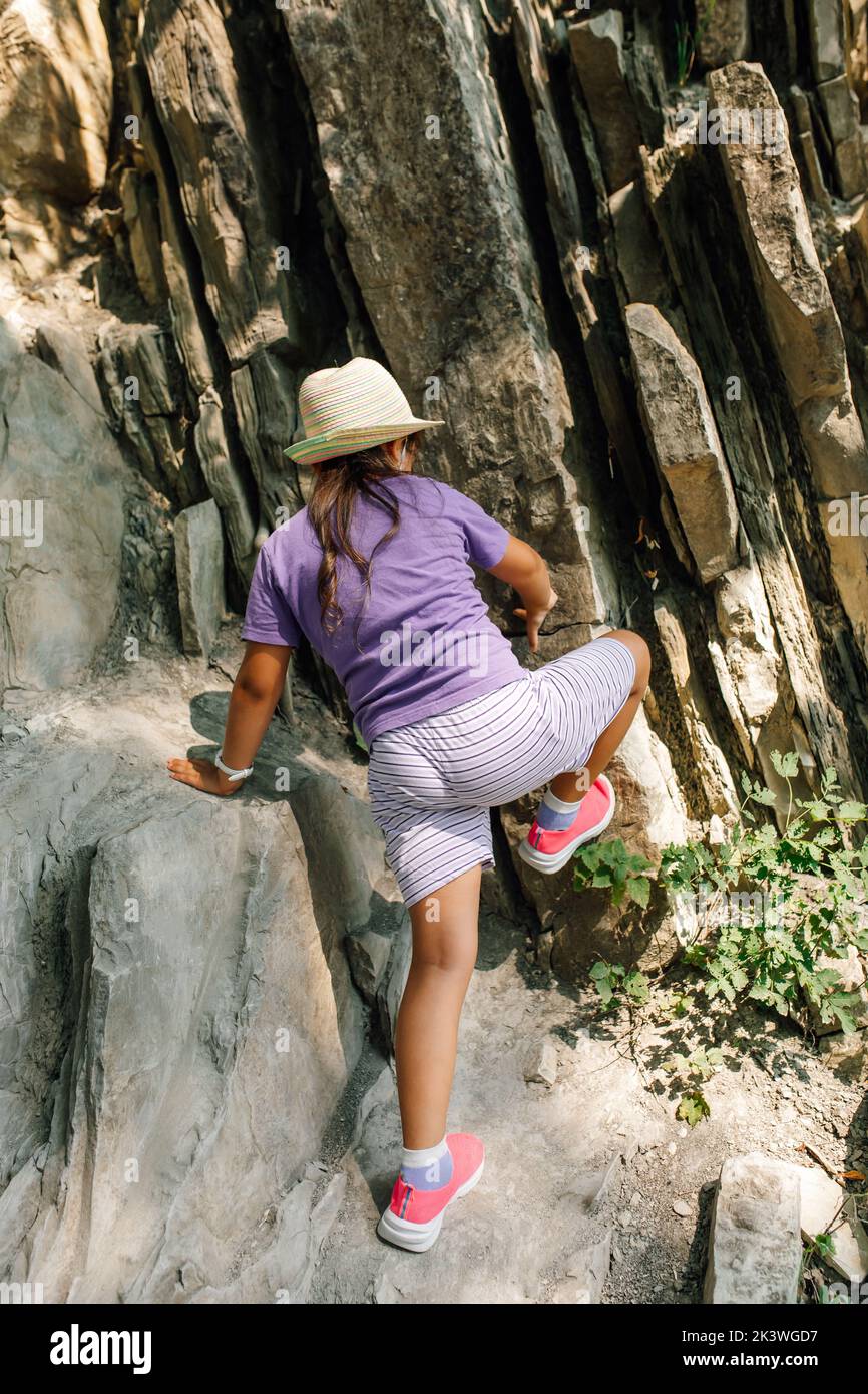 Child in panama hat singly climb up rock in mountains back view. Little girl in sports clothes and footwear go down dangerous route. Tourism Stock Photo