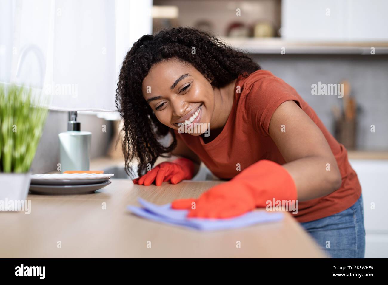 Smiling pretty millennial african american woman in rubber gloves dusting table with dishes Stock Photo
