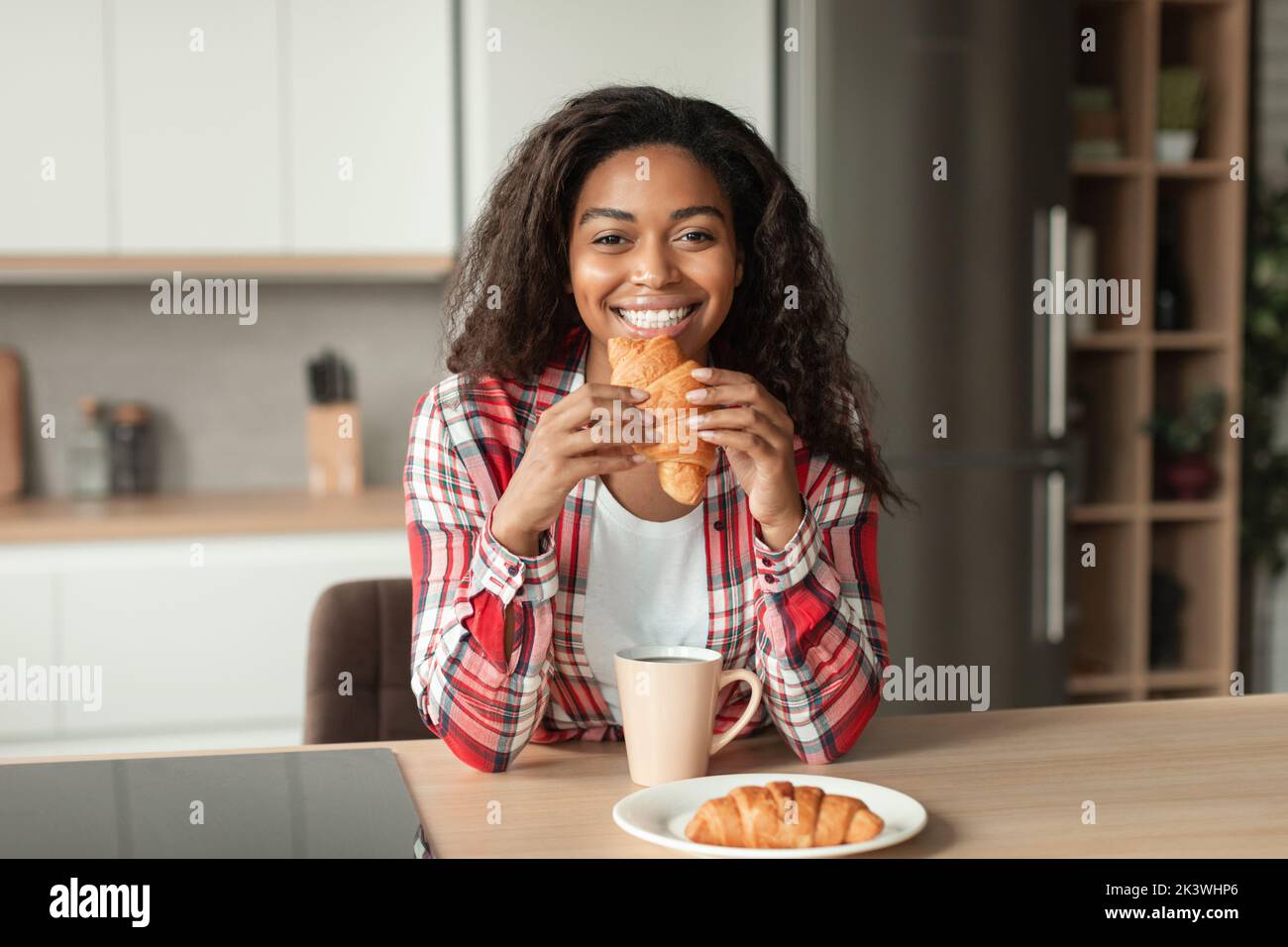 Smiling millennial african american woman with cup enjoy drink, eat croissant in minimalist kitchen interior Stock Photo