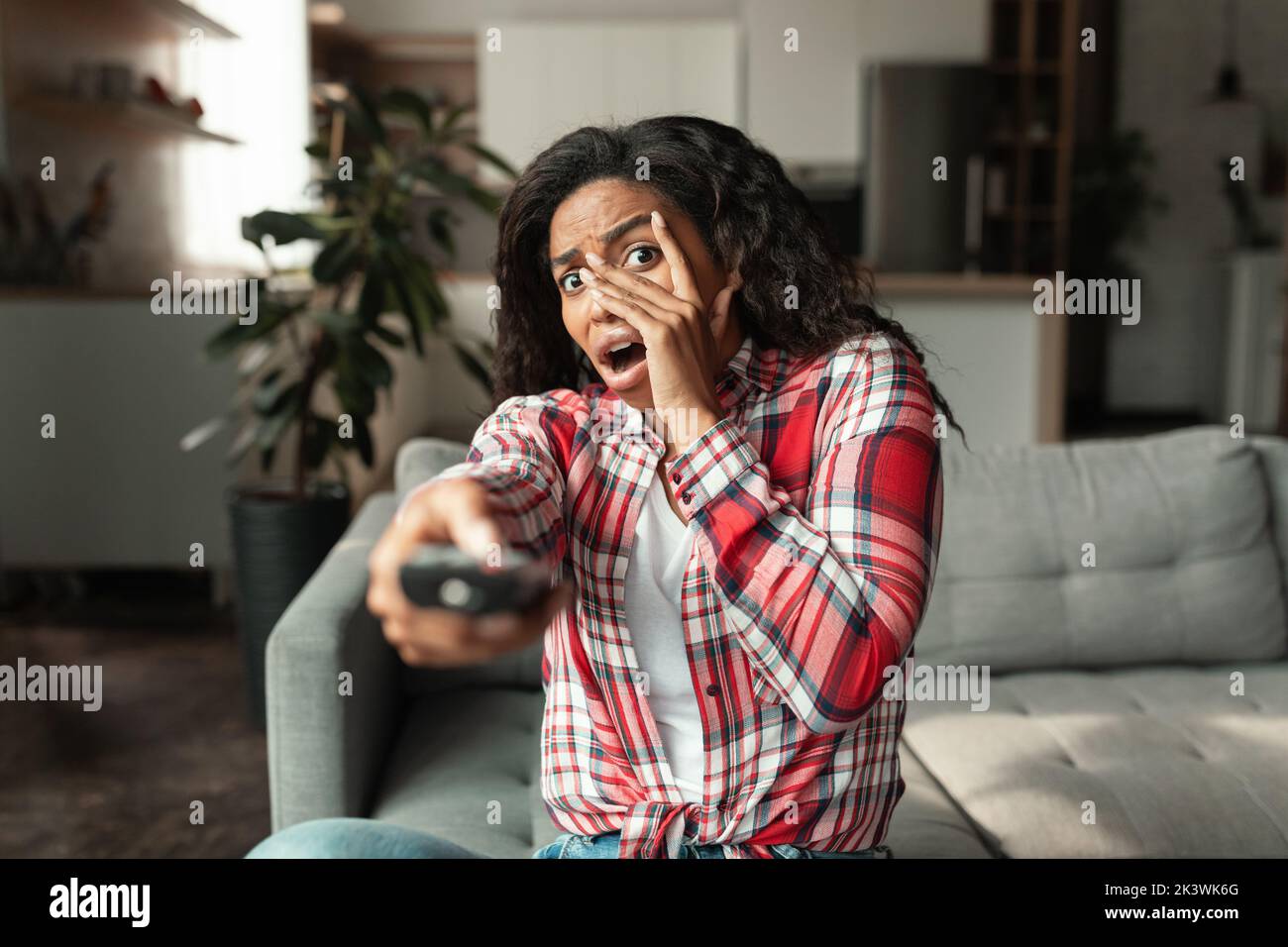 Afraid millennial african american woman with remote control sits on sofa and covers face with hands Stock Photo