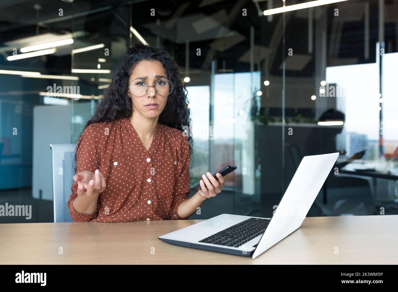 Frustrated and sad business woman with phone in hands looking at camera, latin american woman working with laptop inside modern office building, unclear emotional state of female employee. Stock Photo