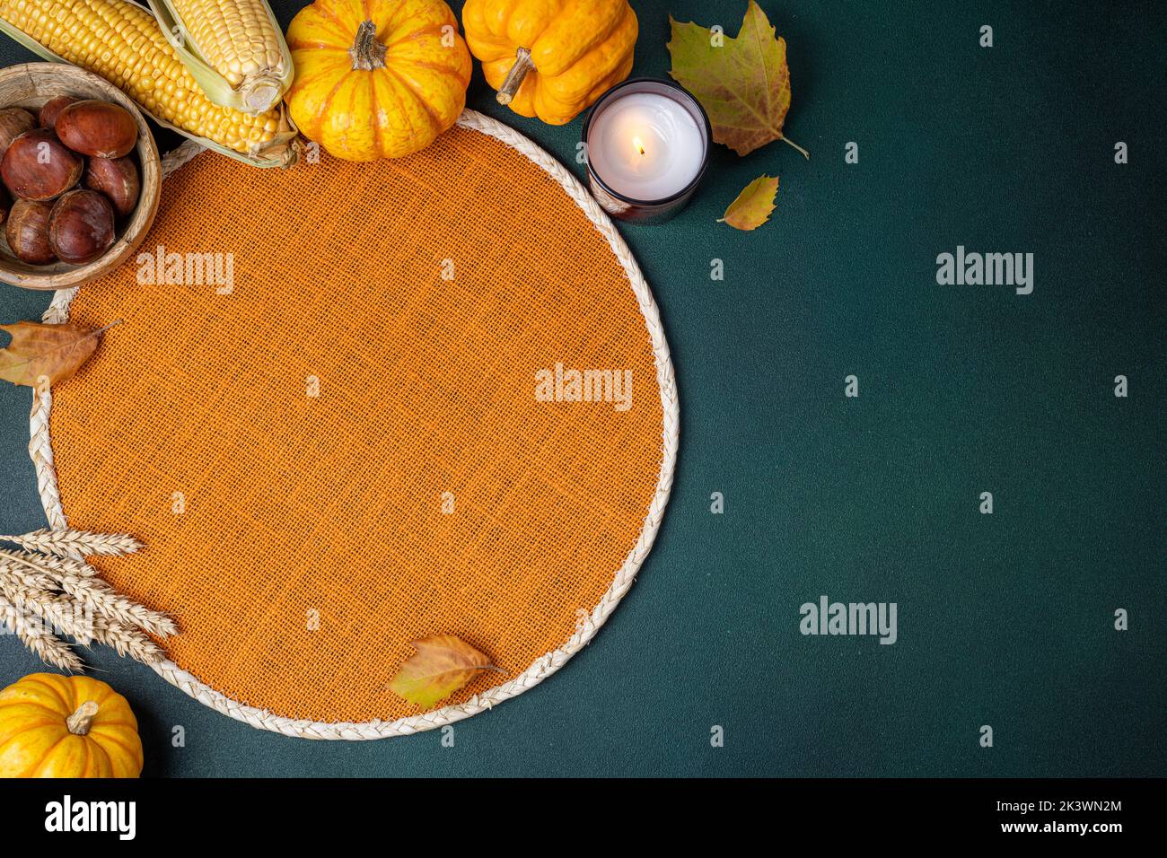 Top view of wicker orange place mat, pumpkins, harvest corn, chestnut, autumn leaves, candle on dark table. Restaurant menu. Table setting on autumn b Stock Photo