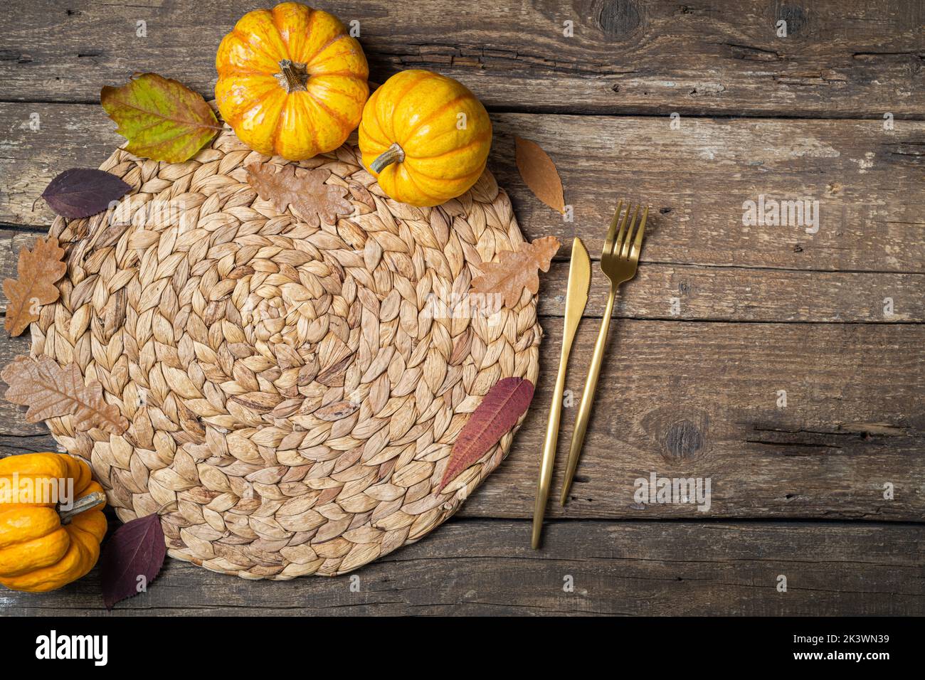 Restaurant menu. Top view of wicker straw place mat, pumpkins, autumn leaves and cutlery on rustic wooden table. Table setting on autumn background. C Stock Photo