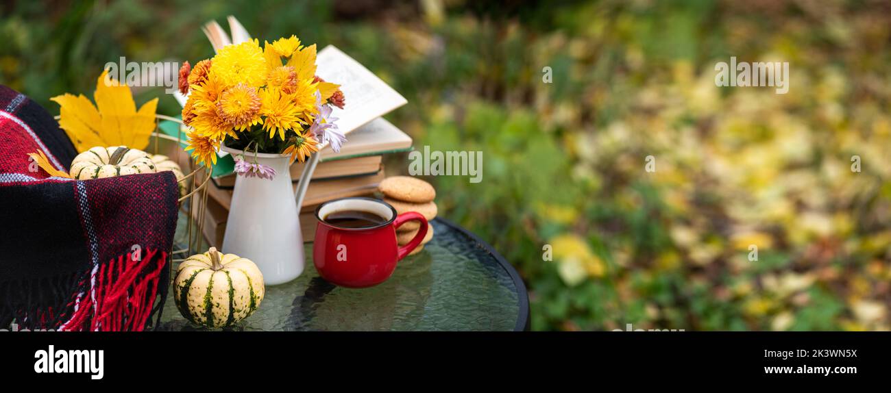 Autumn composition in autumn garden. Warm woolen red blanket, pumpkin, coffee cup, cookies, books, decorations, autumn leaves on table. Fall backgroun Stock Photo