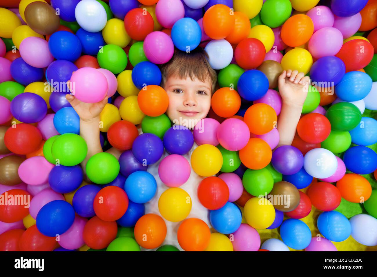 Smile kid lying colorful plastic balls pool. Playroom kids ball pit. Colorful balls dry pool kindergarten playground child indoor play area. Caucasian Stock Photo
