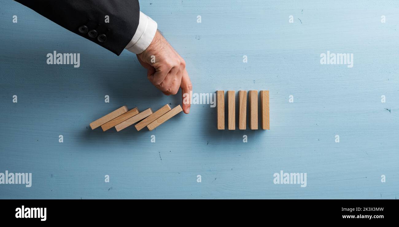 Hand of a businessman interrupting collapsing dominos with his finger in a conceptual image of business crisis management. Over pastel blue background Stock Photo