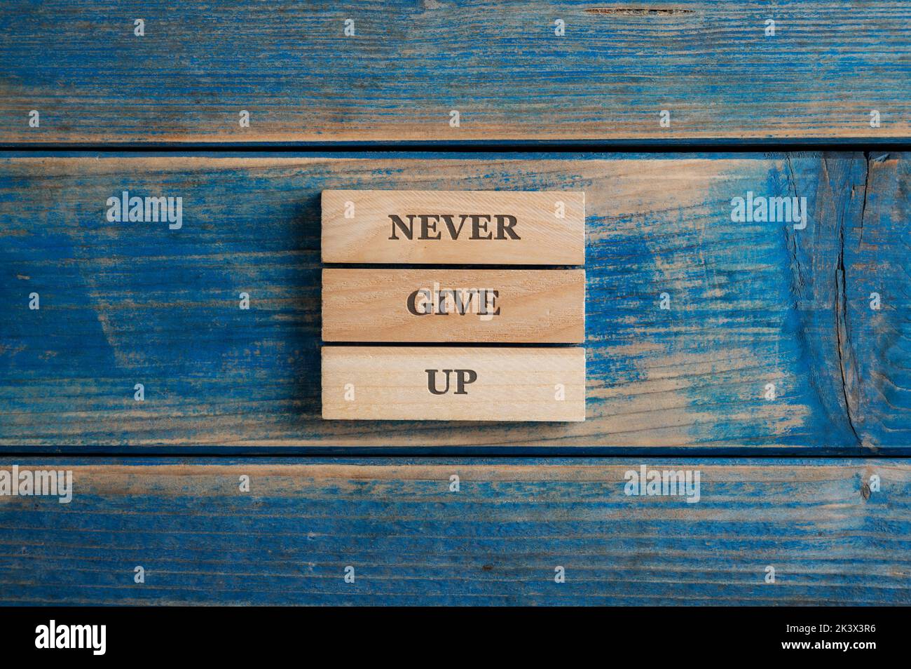 Never give up sign spelled on a stack of three woden pegs placed over textured blue wooden background. Stock Photo
