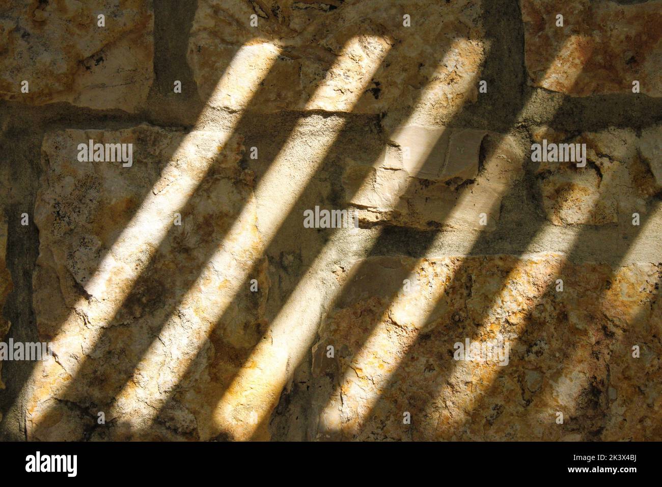 Background - Ancient rough rock wall in deep shadow with diagonal bars of bright light. Stock Photo