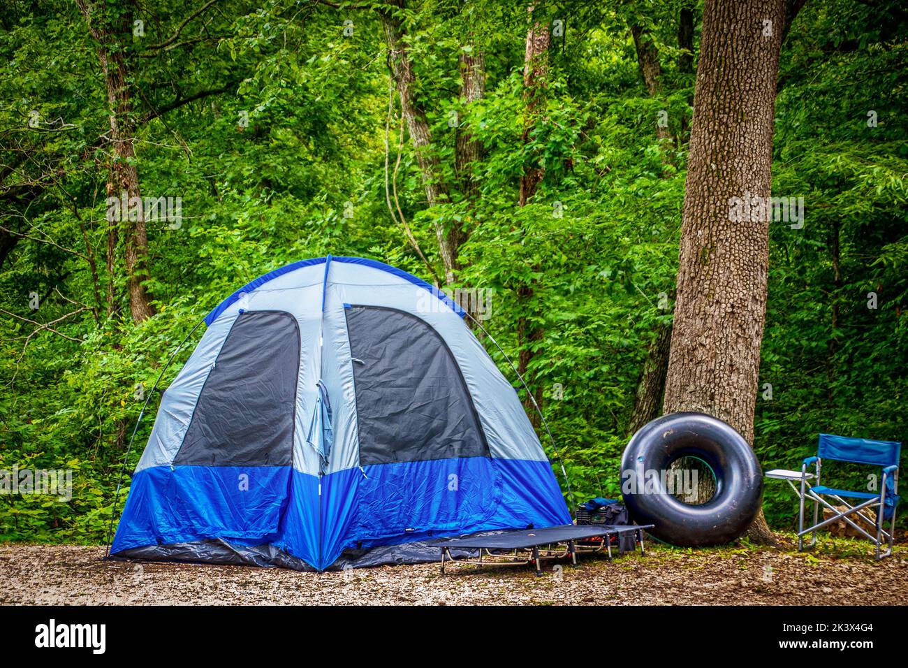 Camping in the woods - Basic blue tent with simple bed outside and tube floaty proped against tree by camp chair - lush foliage background Stock Photo