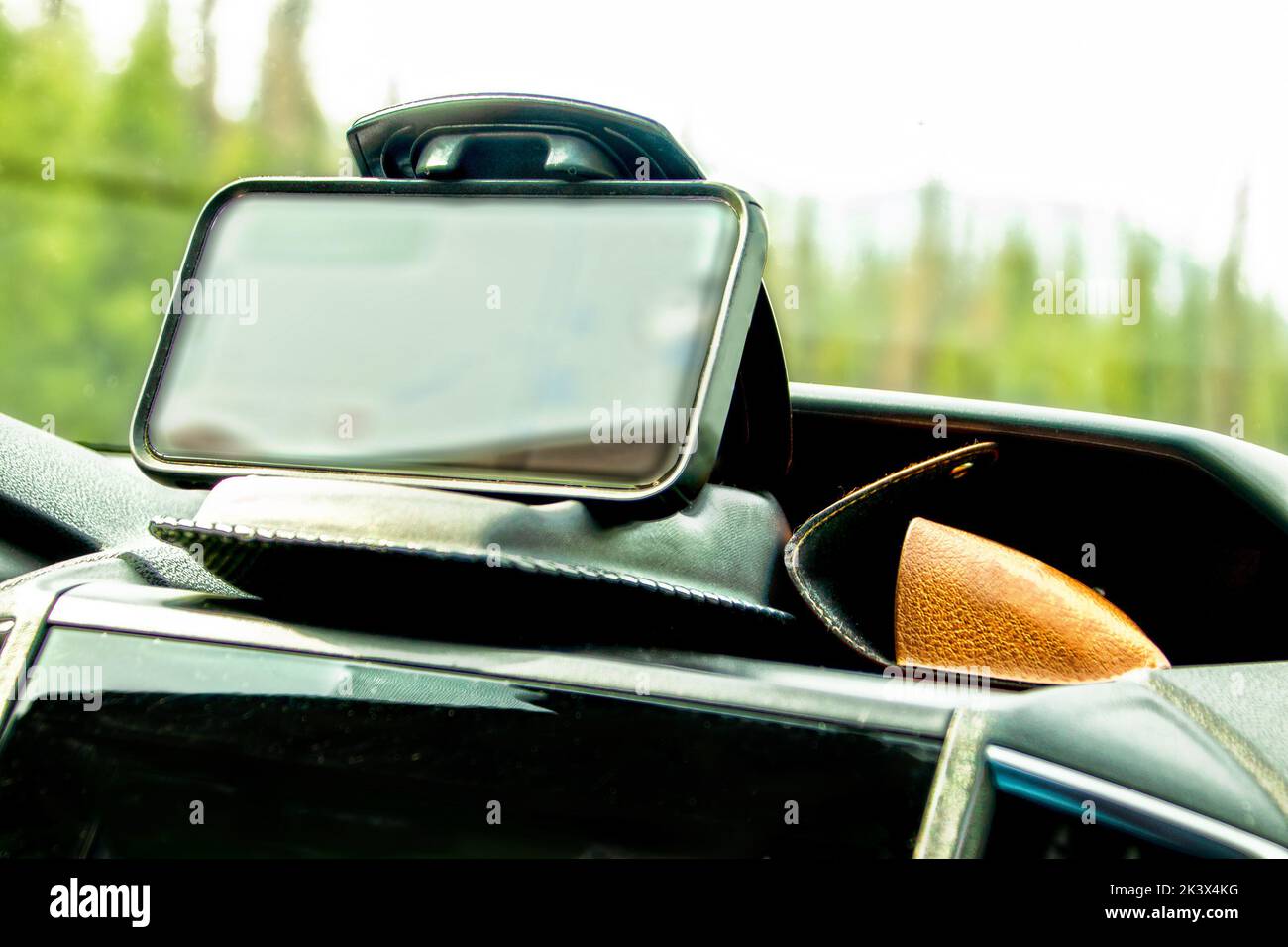 Mobile phone in a weighted clamshell clamp and sunglasses case on dashboard of vehicle with blurred trees in windshield - Soft focus Stock Photo