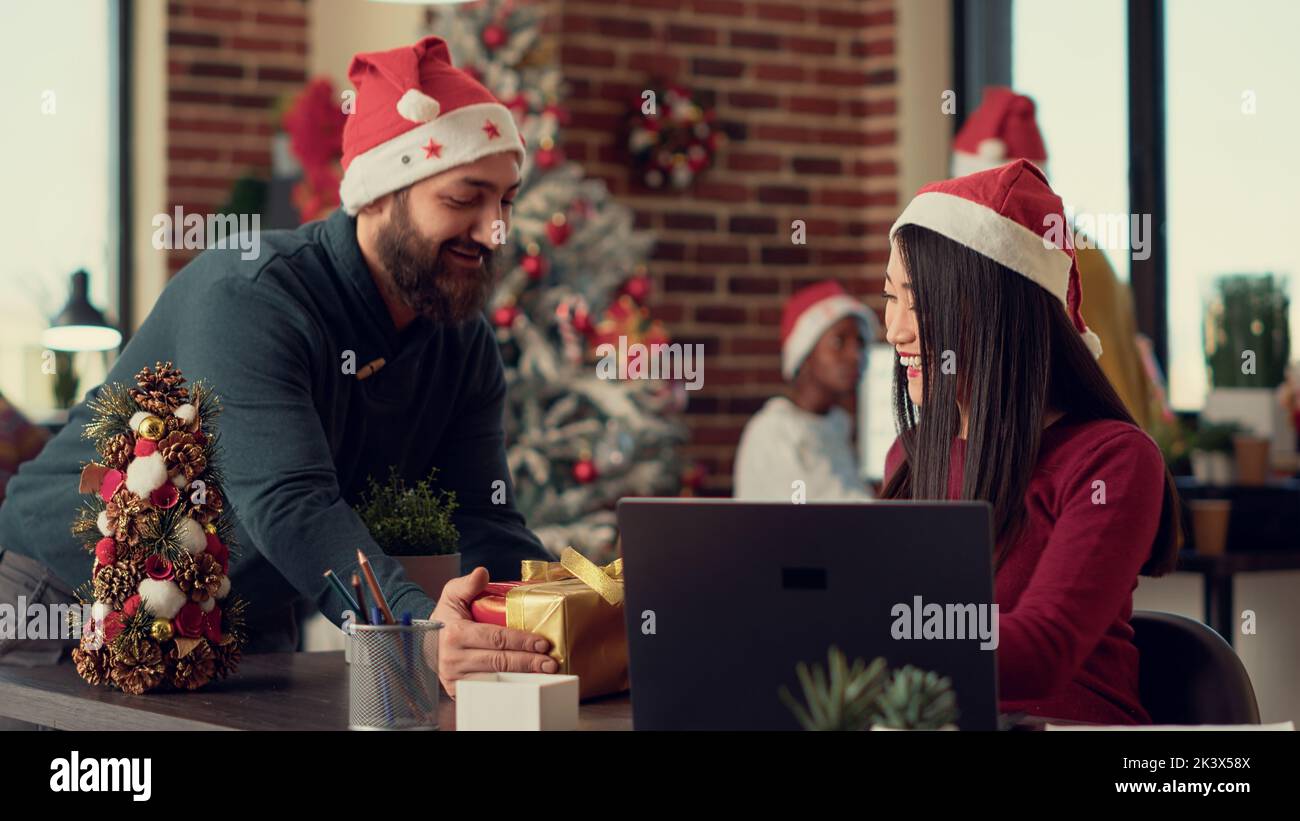 Asian woman receiving christmas present from colleague during holiday season in festive office. Cheerful people exchanging gifts and celebrating xmas eve in space decorated with ornaments. Stock Photo