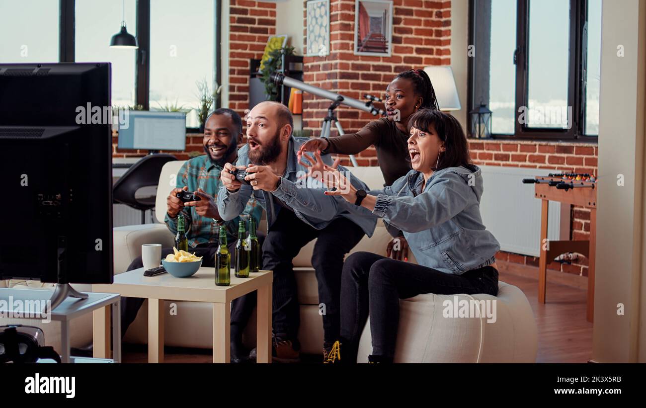 Diverse people winning video games on console, playing online competition together on television. Celebrating challenge win and gaming victory at fun party with beer and snacks. Stock Photo