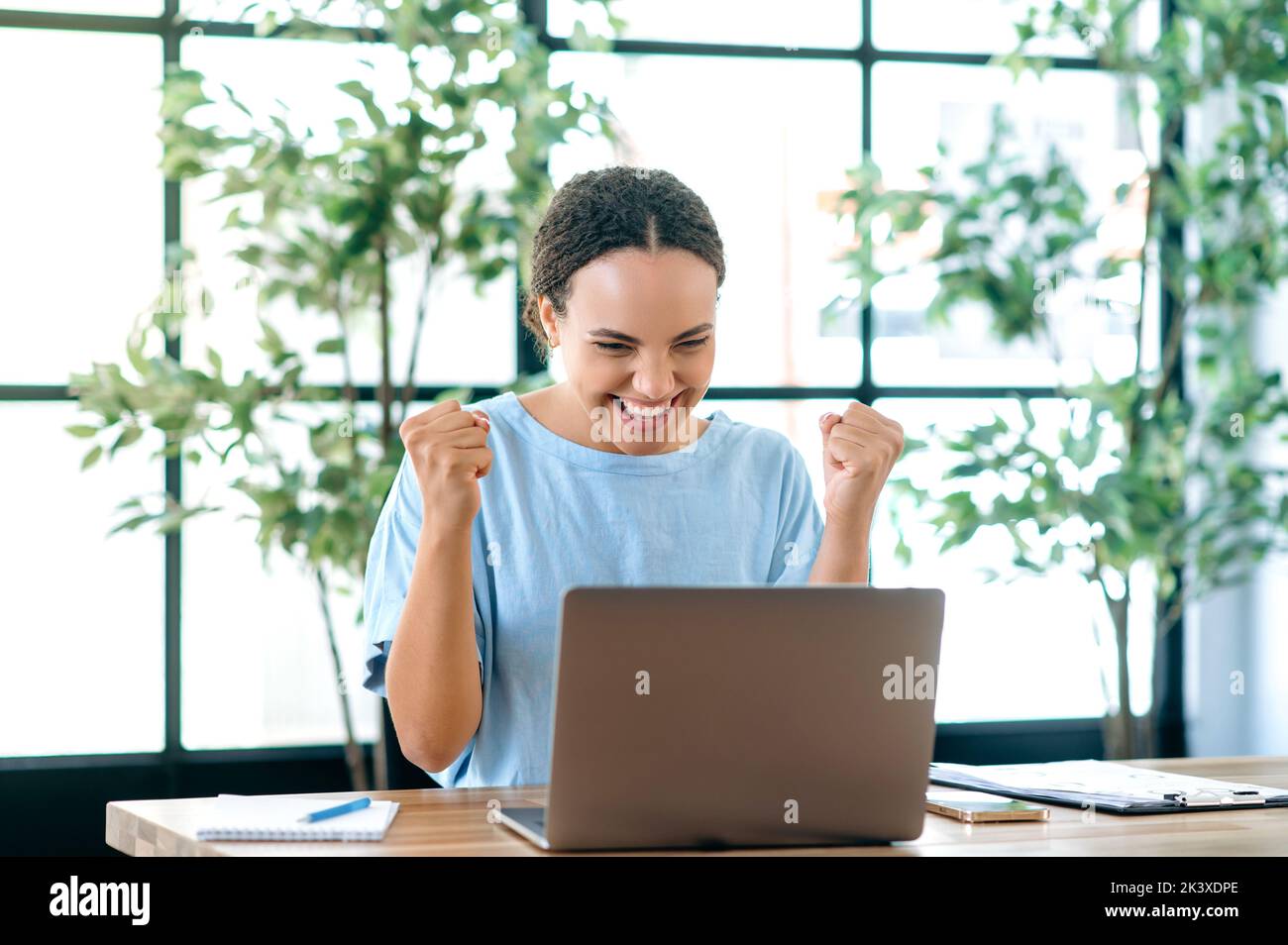 Win, success. Cheerful happy brazilian or hispanic young business woman, sits at a desk with a laptop in modern office, rejoicing in success, big profit, celebrate deal, gesturing with fists, smiling Stock Photo