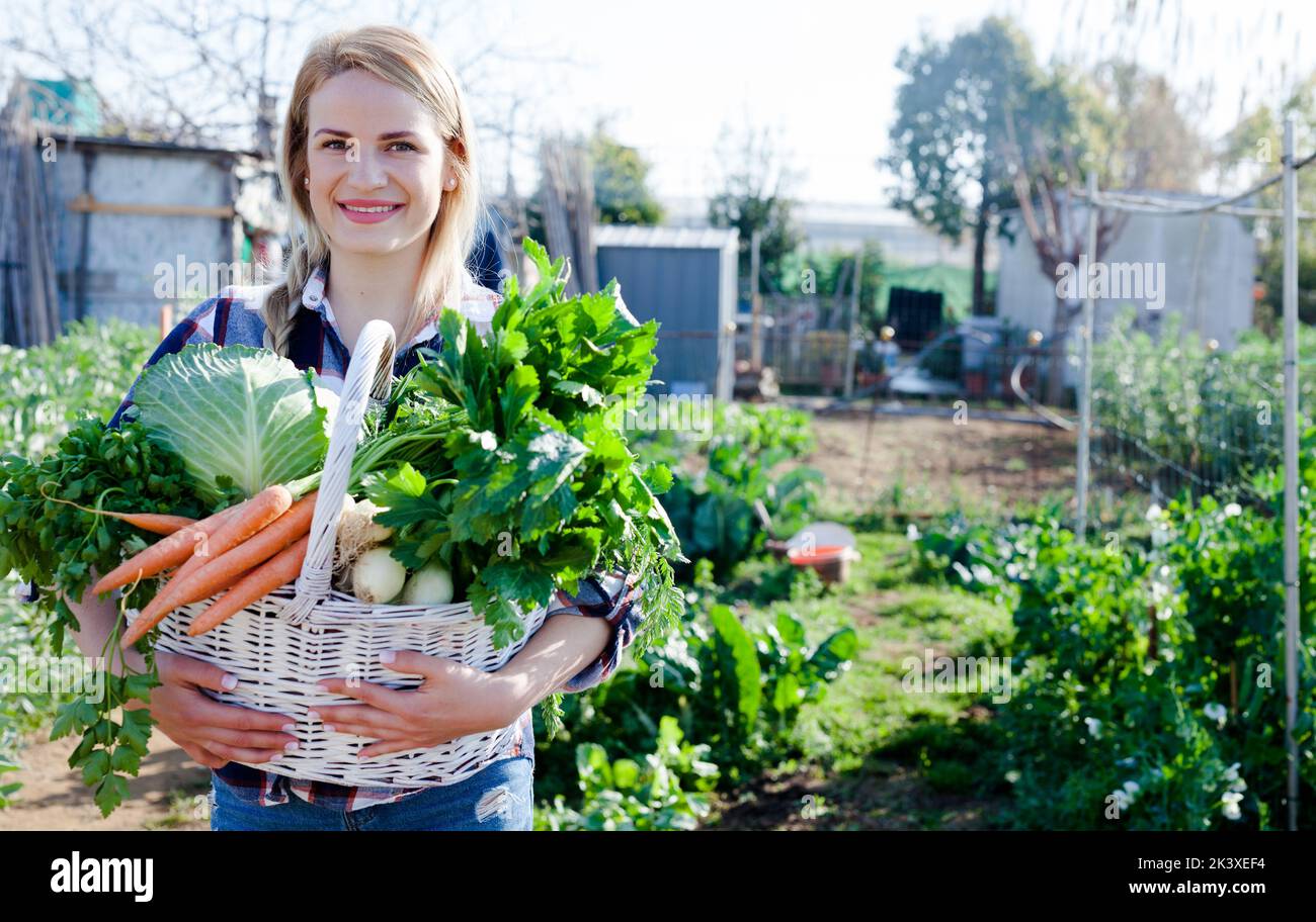 Young woman gardener holding harvest of vegetables and greens in basket ...