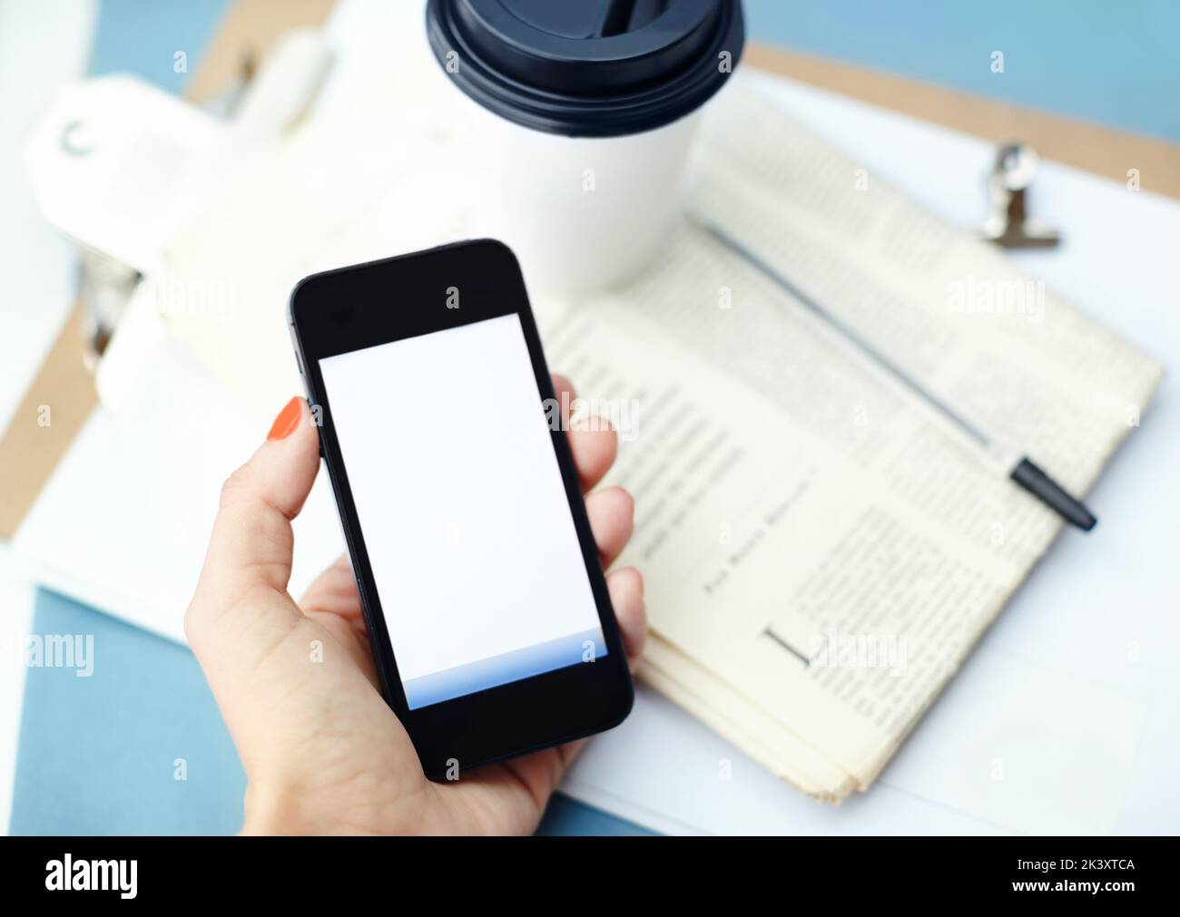 Checking messages between sips of coffee. Hand holding a smartphone with disposable cup, pen, binder clip, newspaper and clipboard. Stock Photo