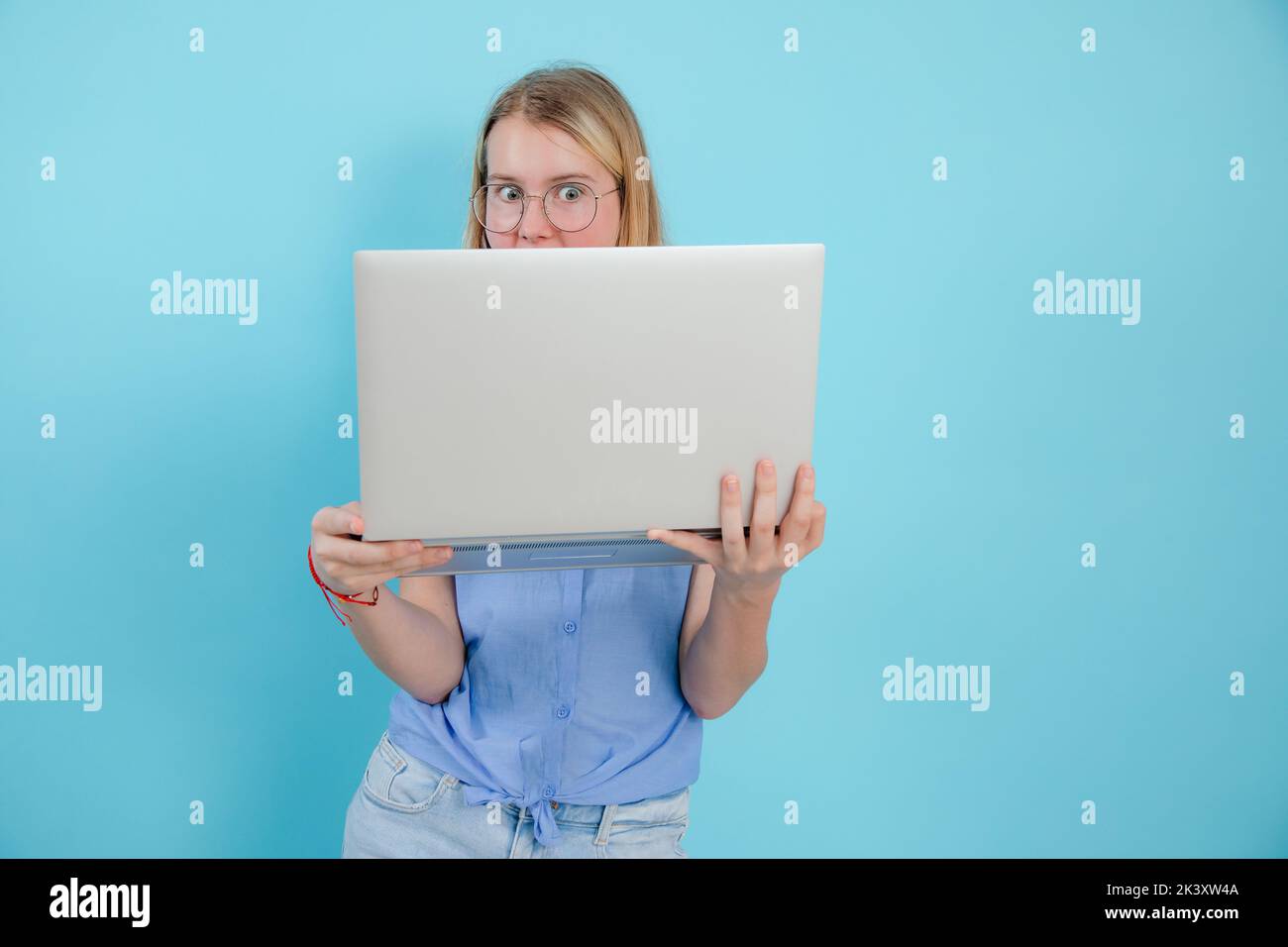 Portrait of shocked teenage girl with long hair wearing blue sleeveless shirt, holding laptop on blue background. Stock Photo