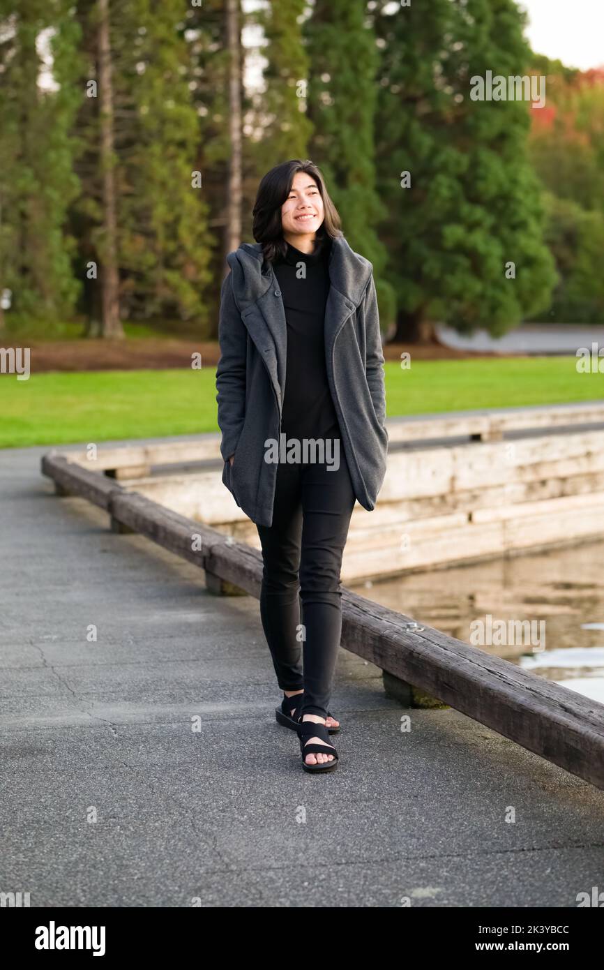 Smiling  teen girl in gray jacket walking along path by lake at sunset Stock Photo