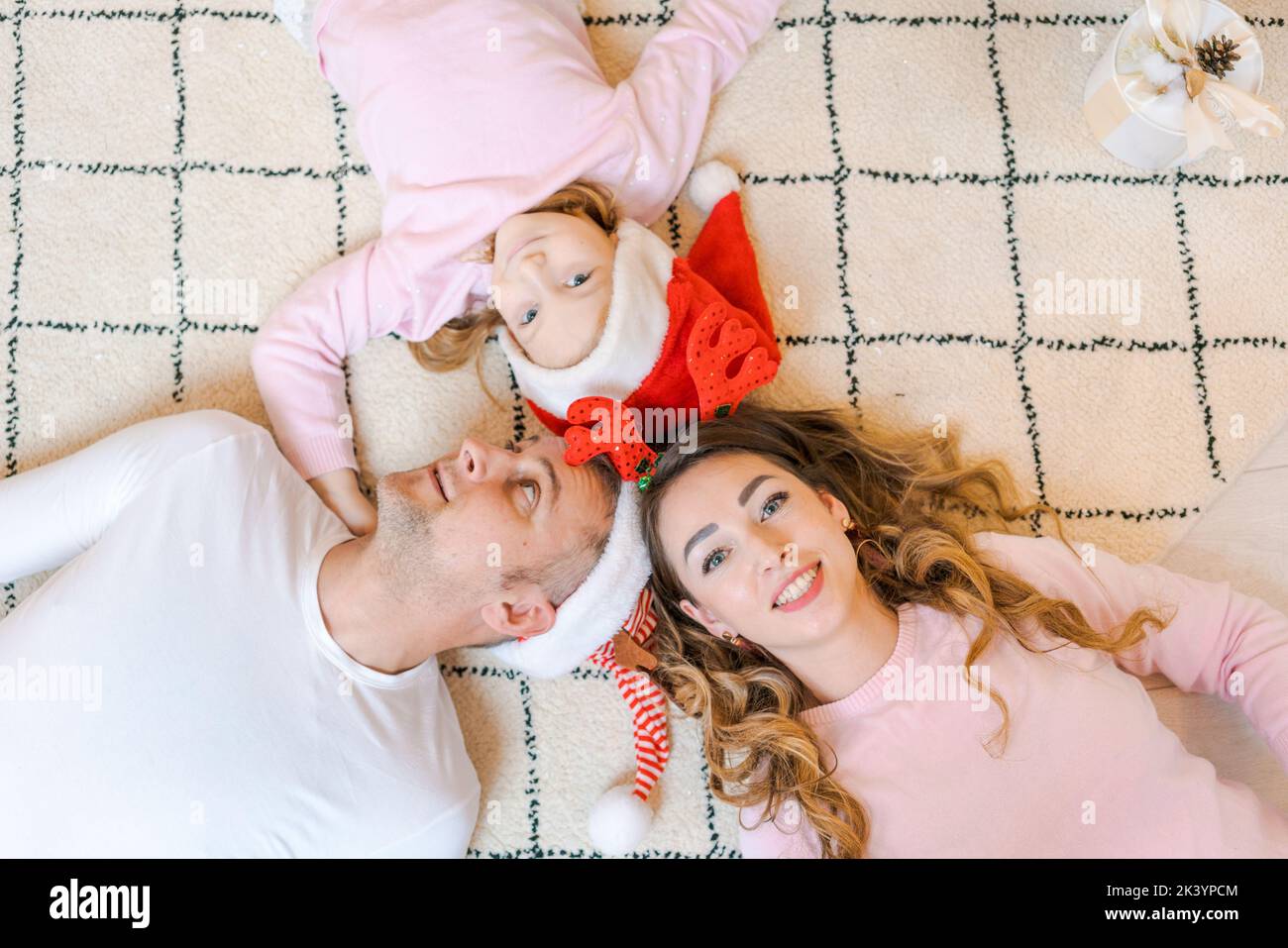 Family in Santa hats with their daughter, in light, cozy clothes, lying on floor in a decorated house, not far from the Christmas tree. Merry Christmas and Happy New Year concept Stock Photo