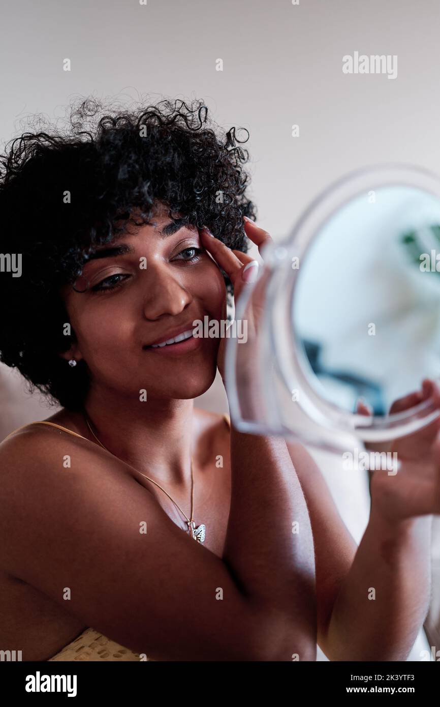 Confident young transgender woman with curly hair smiling while looking herself in a small mirror. Stock Photo