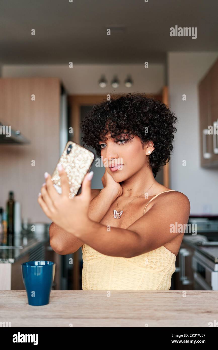 Confident young transgender woman taking selfies with a smartphone while sitting in the kitchen at home. Stock Photo
