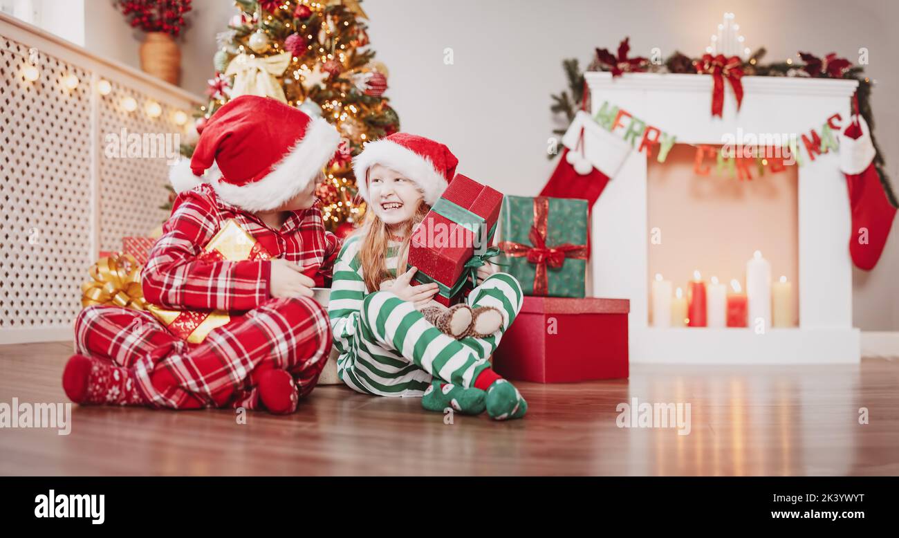 Brother and sister sitting on the floor indoors with Christmas gift boxes in their hands. Concept of the holiday eve and New Year. Stock Photo