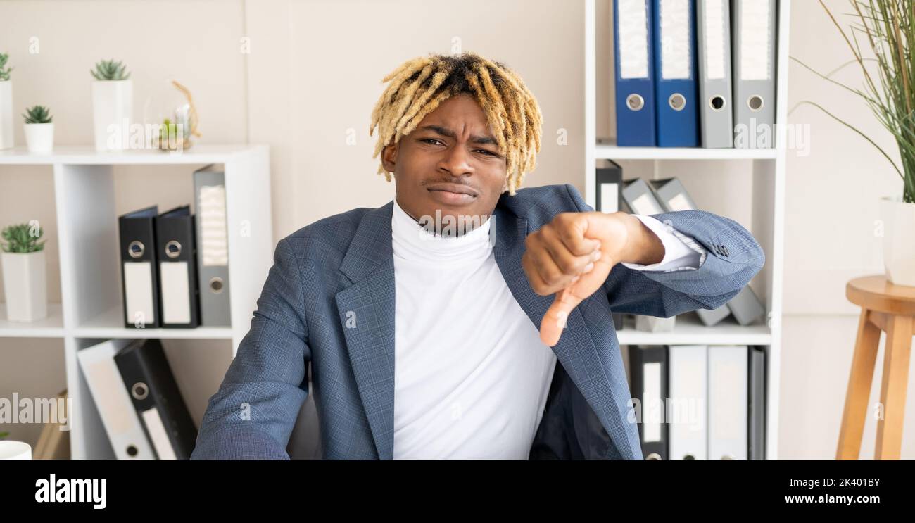 Bad job. Displeased black man. Negative result. Disappointed male worker sitting office desk showing thumb down dislike gesture in light room interior Stock Photo