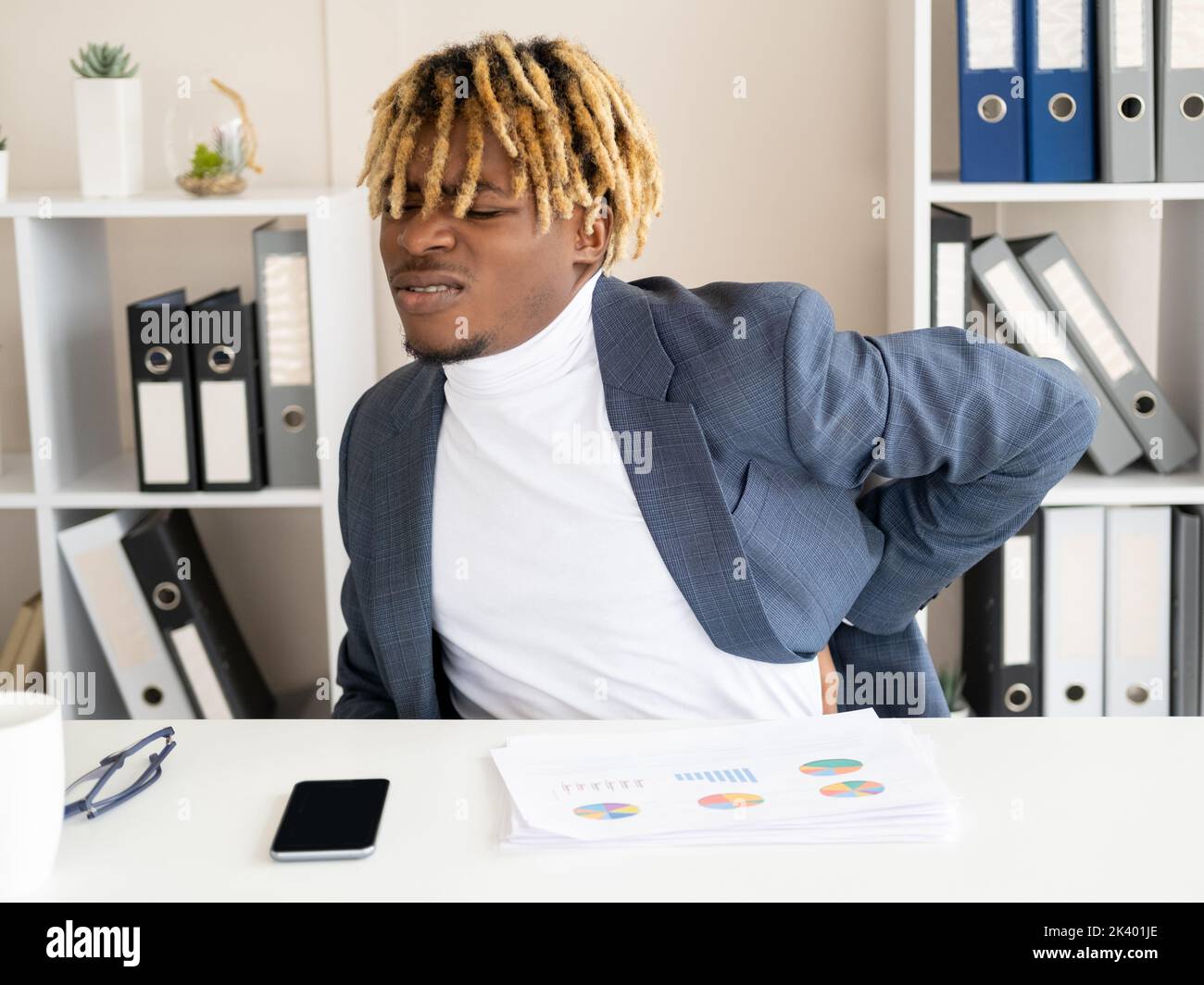 Sitting office work. Sick black man. Back pain. Exhausted guy holding hand on neck grimacing from ache sitting desk light room interior. Stock Photo