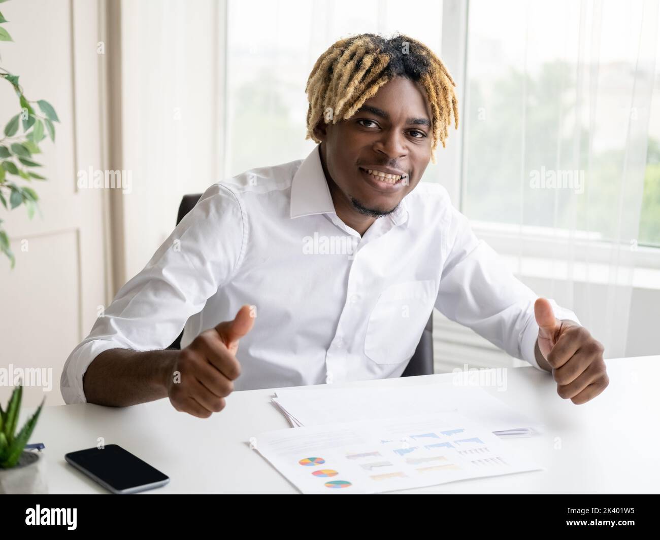 Successful result. Happy black man. Enjoying work. Positive smart casual guy sitting office desk showing likes gesture in light room interior. Stock Photo