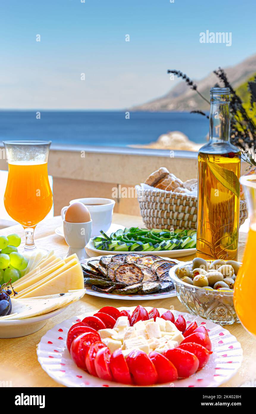 Home-made fresh summer breakfast with orange juice, cherry tomatoes, cheese, olives, cucumber, eggs a cup of coffee on the balcony with sea view.  Bac Stock Photo