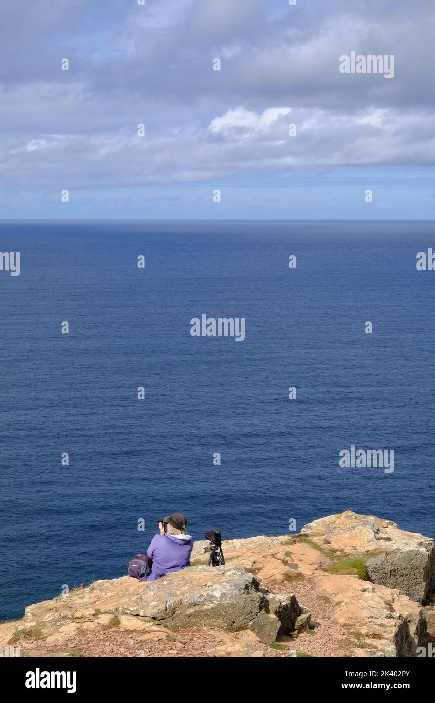 A marine wildlife watcher sits on St Agnes Head looking out to sea with binoculars and telescope. Stock Photo