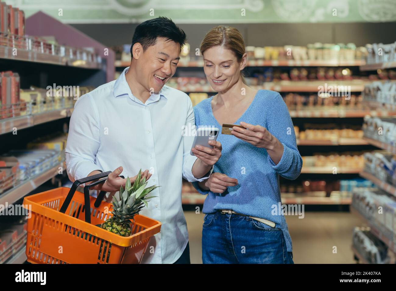 Family shopping. Young beautiful interracial couple shopping in a supermarket. Happy Asian woman and man holding credit card and phone, paying cashless, making online order. Stock Photo