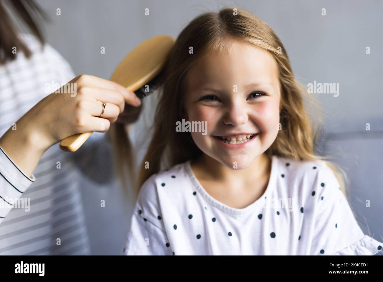 Charming little girl is looking at camera and smiling while her beautiful mother is combing daughter's hair Stock Photo