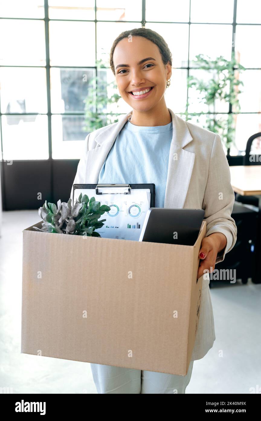 New job. Vertical photo of joyful successful motivated mixed race woman, stand in her new working office, holding a cardboard box with work attributes in her hands, looks at camera, smiling happily Stock Photo