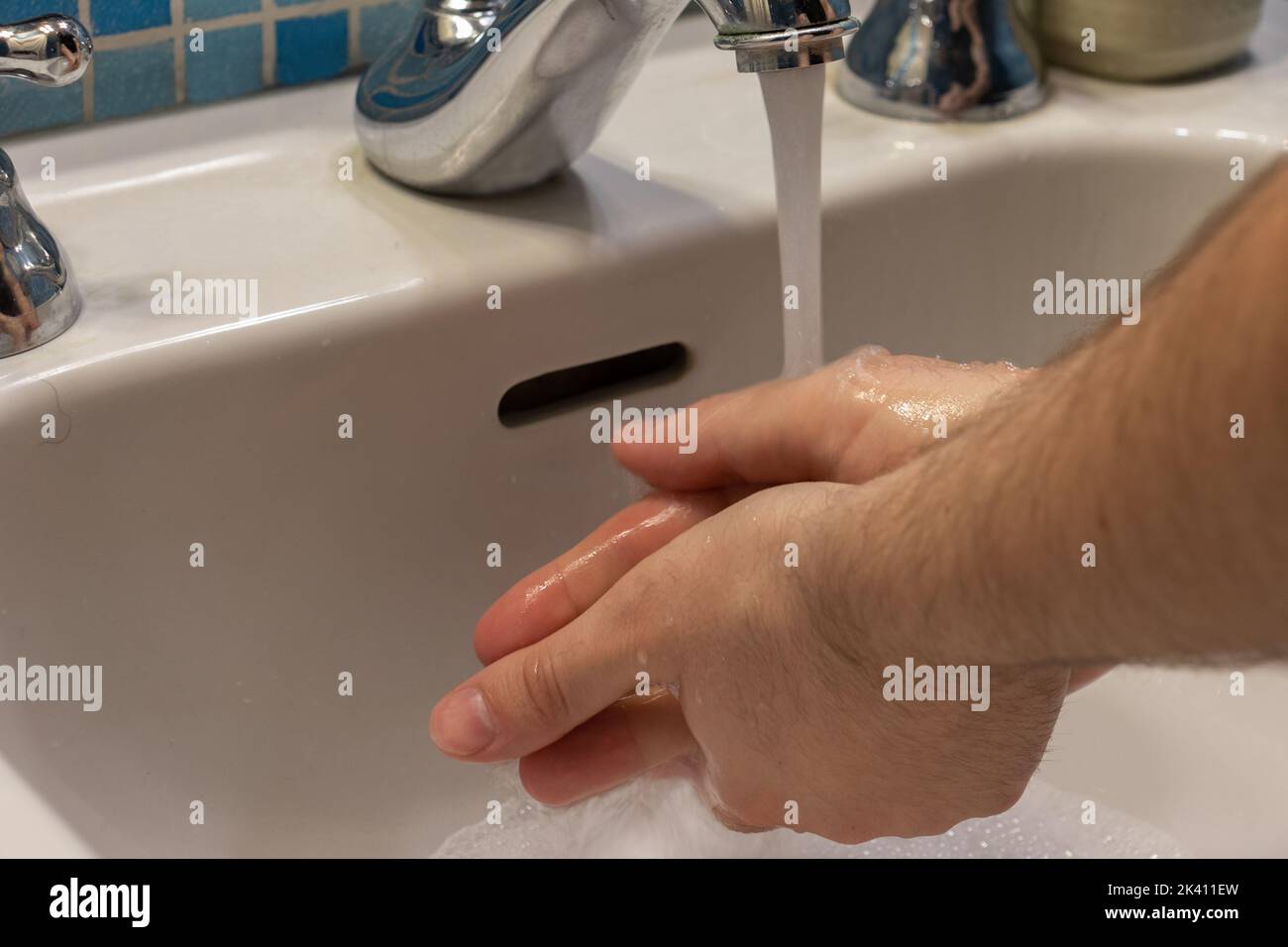 Caucasian man washing his hands with soap in an act of hygiene in a white sink with blue-toned tiles Stock Photo
