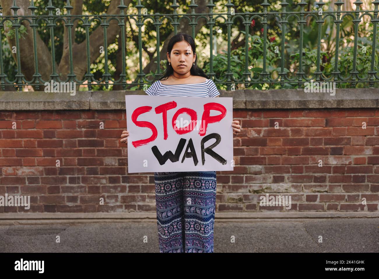 Young teenage girl looking at the camera while holding an anti-war poster. Young peace activist protesting against war and violence. Stock Photo