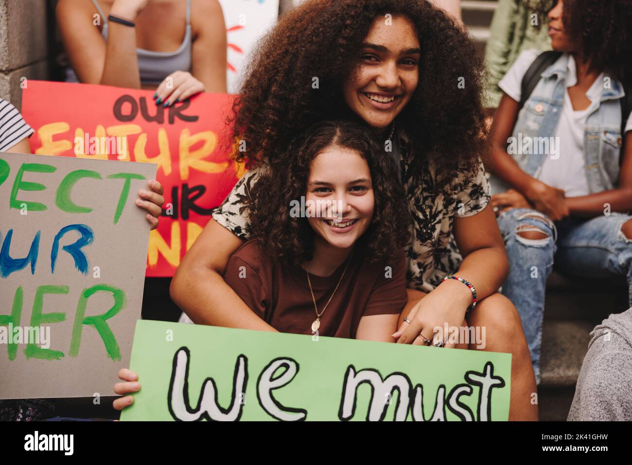 Cheerful teenage girls smiling at the camera while sitting with a crowd of climate activists. Group of multicultural protestors striking for climate j Stock Photo