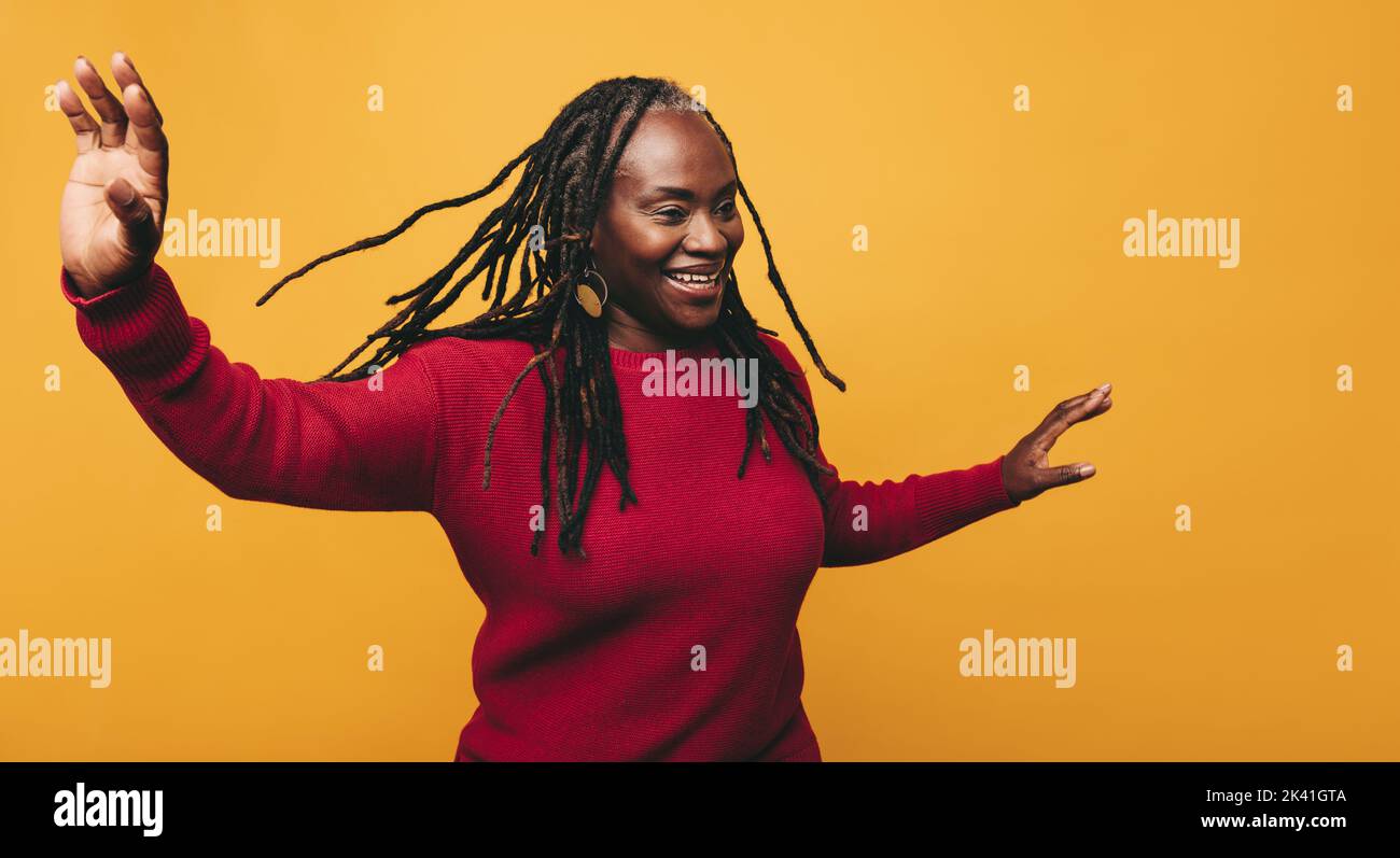 Happy mature woman dancing and having fun while standing against a yellow background. Cheerful black woman with dreadlocks laughing joyfully in a stud Stock Photo
