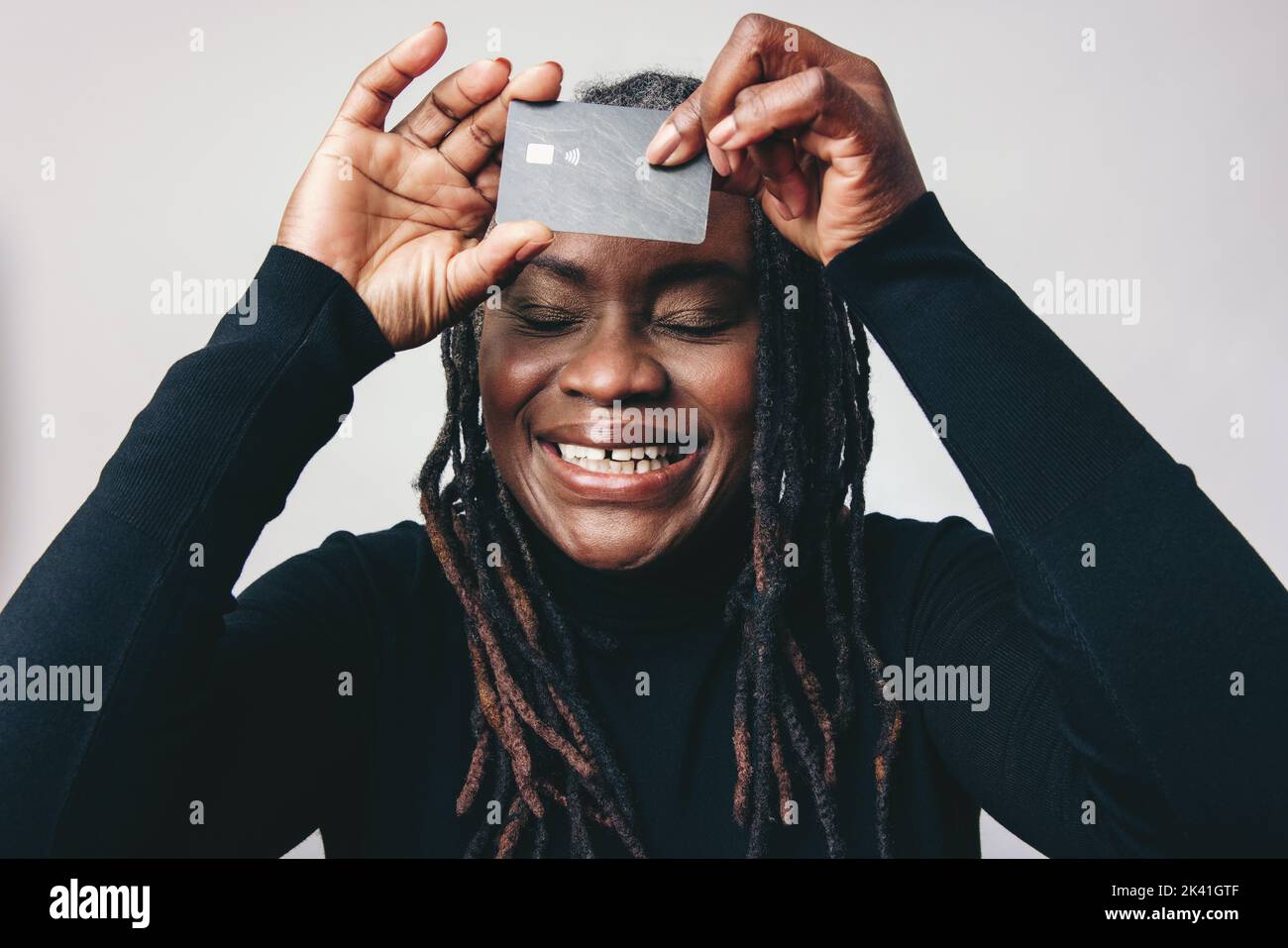 Mature woman with dreadlocks smiling happily while holding a credit card over in front of her forehead. Cheerful middle-aged woman recommending electr Stock Photo