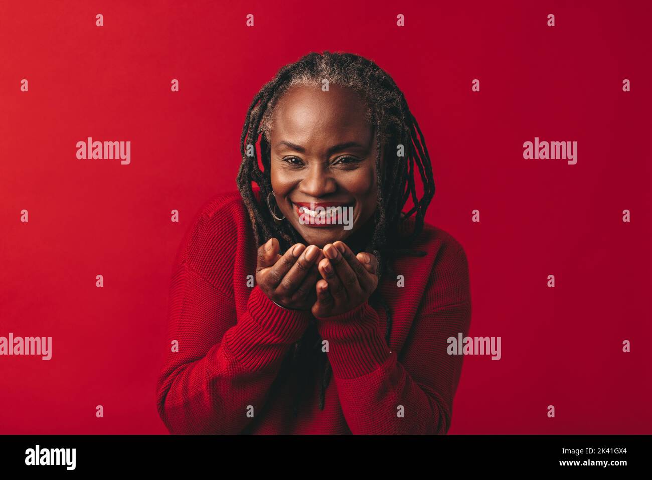 Mature woman smiling and blowing air confetti while standing against a red background. Happy middle-aged woman with dreadlocks having fun in a studio. Stock Photo