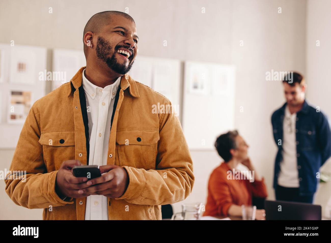 Happy businessman with a septum ring looking away while holding a smartphone. Cheerful young businessman working in a creative and modern workplace wi Stock Photo