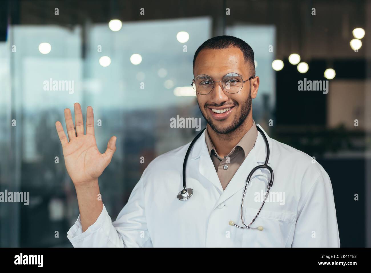 Young african american doctor man wearing glasses wearing medical coat saying hello happy and smiling friendly welcoming gesture doctor working inside clinic. Stock Photo