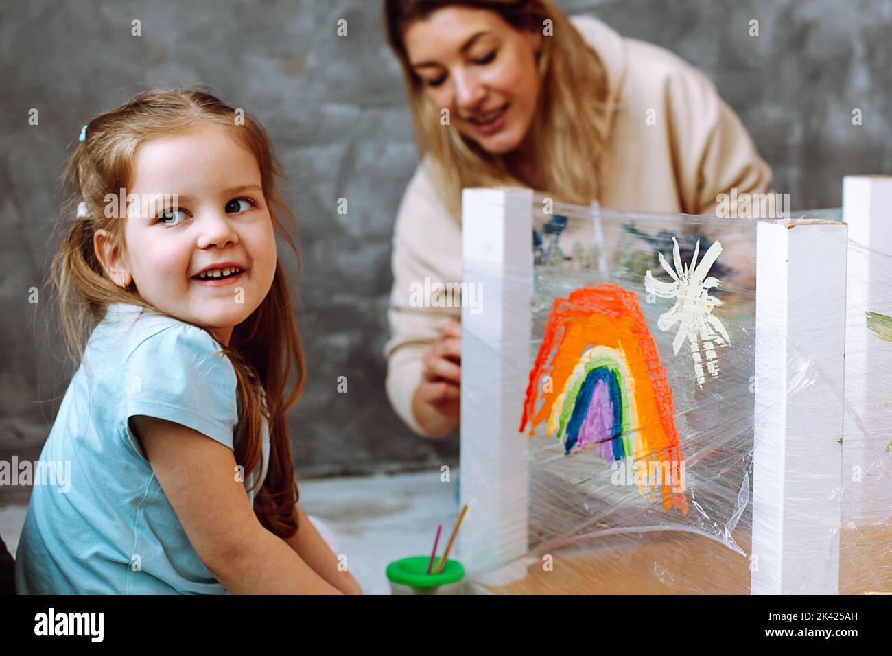 Little smiling blond girl painting rainbow on adhesive tape with teacher. Having fun and learning colors in kindergarten Stock Photo