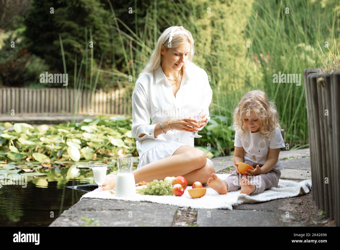 Mother and little blonde daughter with citrus on picnic, have organic fruit and milk lunch in park near lake outdoors Stock Photo