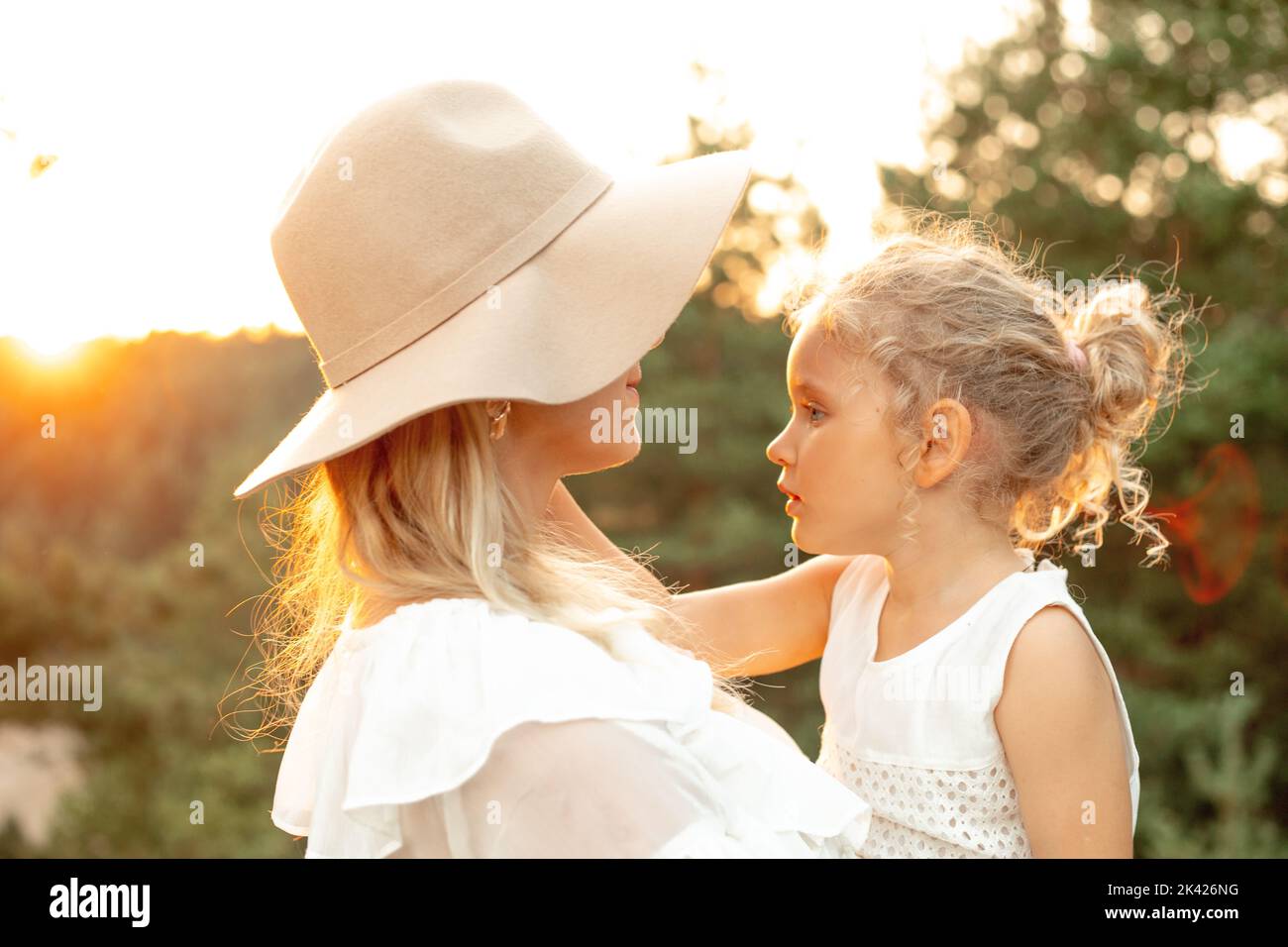Unrecognizable smiling woman in hat embracing little blonde girl, daughter on natural blurred background. Child safety Stock Photo