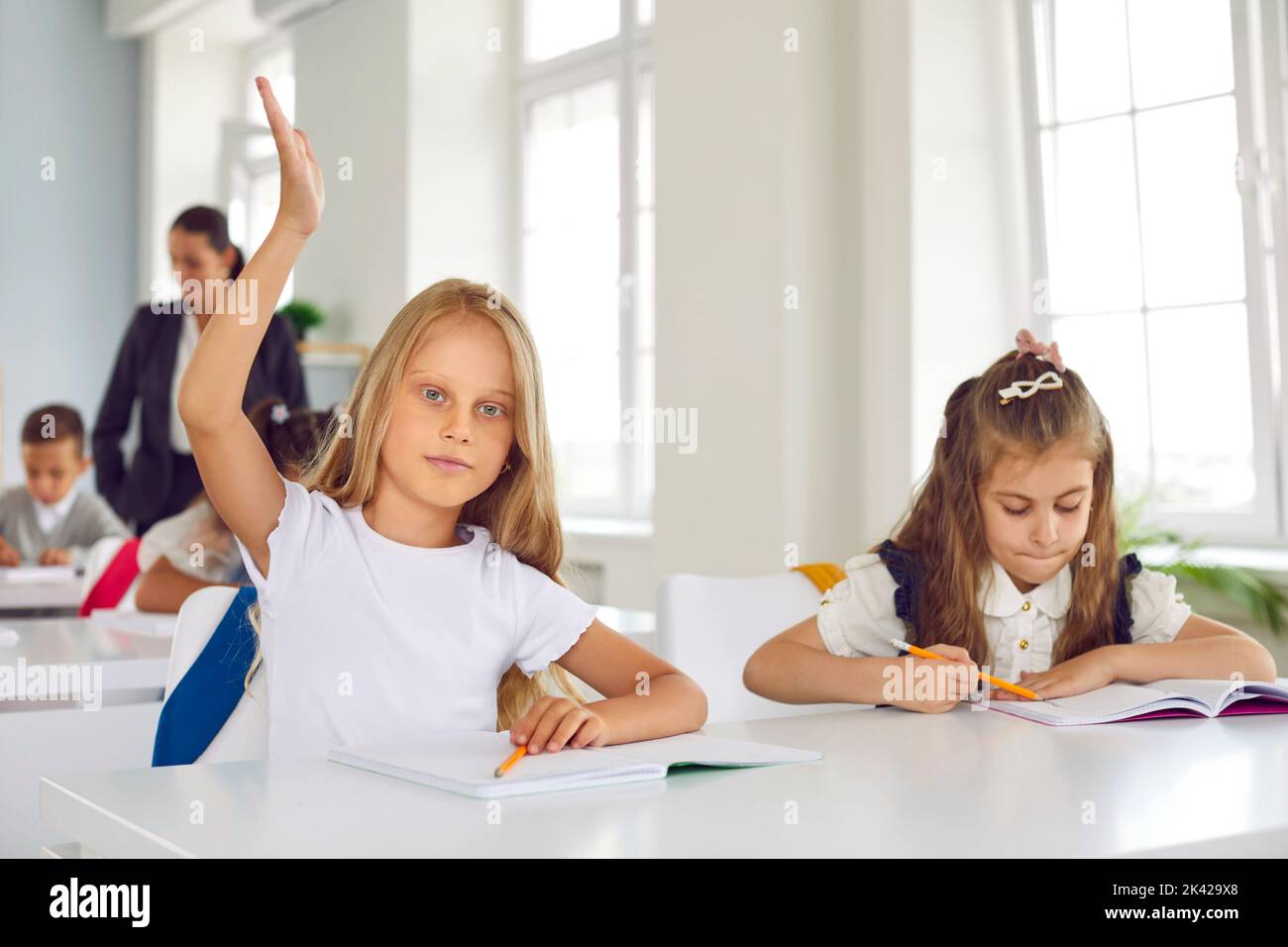Smart little schoolgirl raises her hand during lesson, wanting to answer teacher's question. Stock Photo