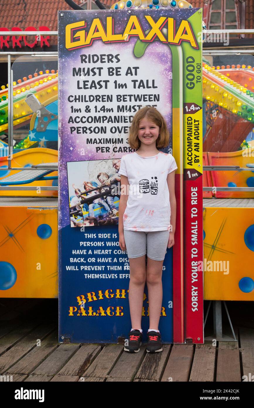 Girl child 1.4 m metres tall girl kid / eight 8 year old girl standing at a height measure measuring scale which indicates she is tall enough to go on a height restricted fairground ride / fair with amusements, attractions, and rides including the Galaxia with its size restriction, at the end of Brighton Palace Pier. UK. (131) Stock Photo