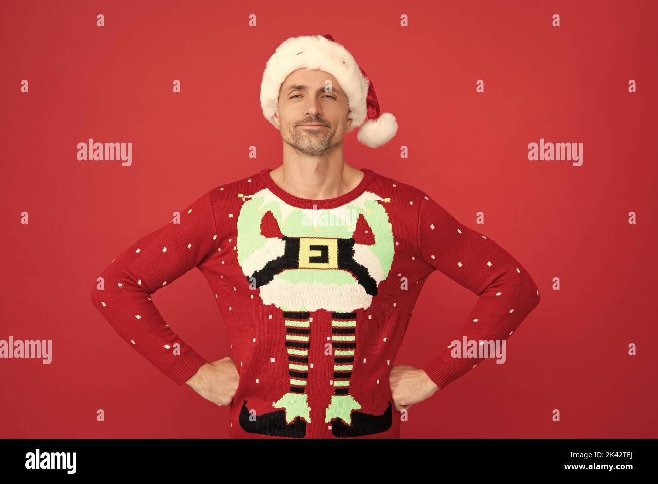Santa Claus elf man in Christmas jumper stand confidently with arms akimbo red background, xmas Stock Photo