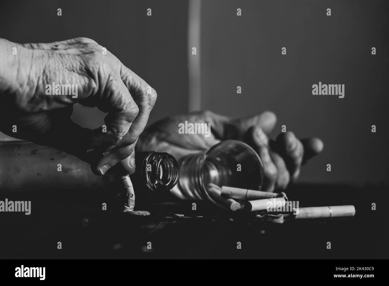Cigarettes a glass and an empty bottle and the hands of an old woman on a burnt black table, female smoking and alcoholism, bad habits Stock Photo