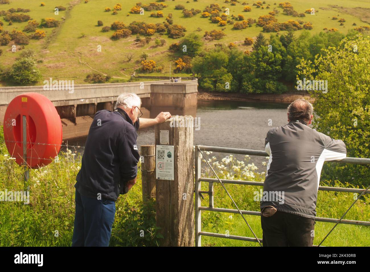 Two men 60+ looking over a gate and fence to the Meldon dam and lake, Devon, England with a life belt to one side on a sunny springtime day Stock Photo