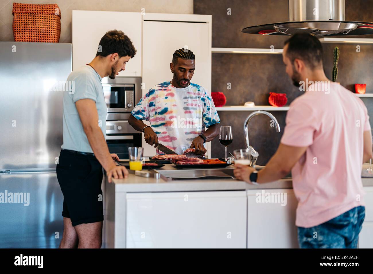 3 friends converse in a kitchen while one cuts meat with a knife Stock Photo